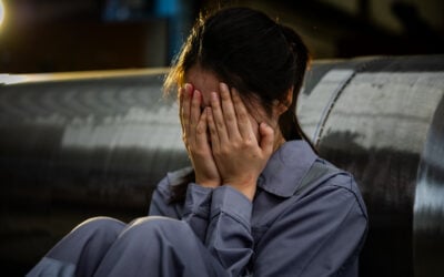 Employee sitting alone after repeated workplace sexual harassment while coworkers laugh in the background, illustrating a hostile work environment and emotional distress under Title VII.